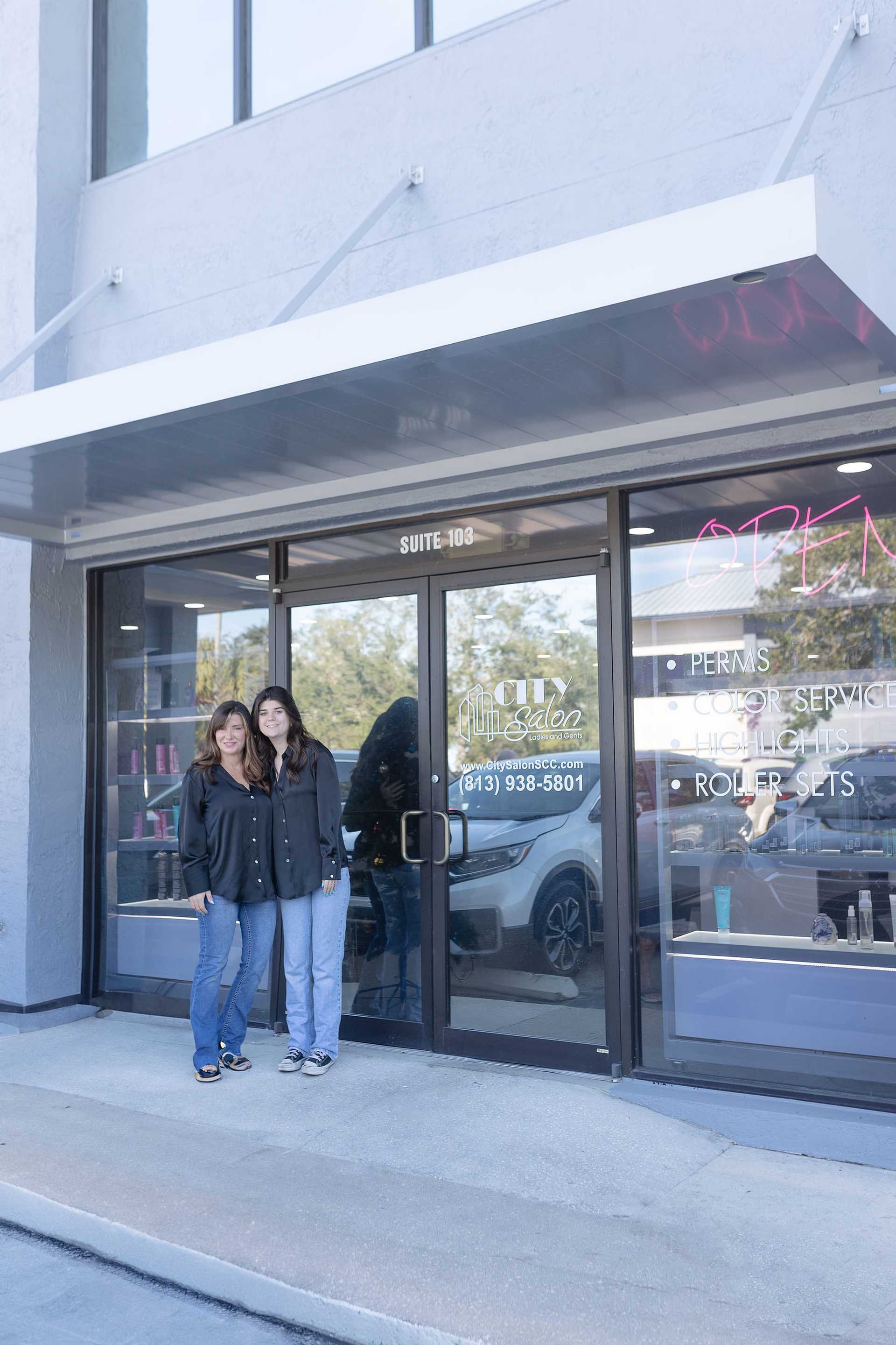 Two people standing in front of a hair salon with "OPEN" sign and service list on the window.